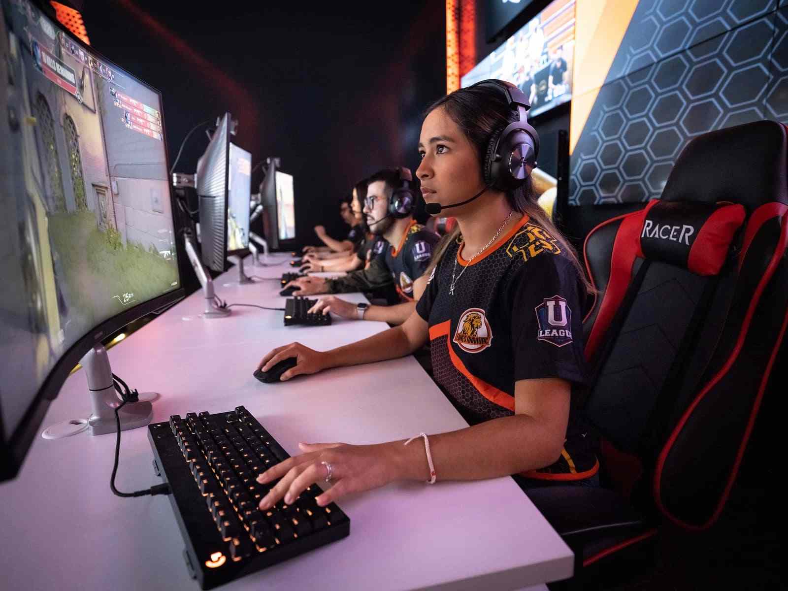 Jugadora con audífonos y camiseta de equipo compite en torneo de eSports usando teclado y monitor gamer.